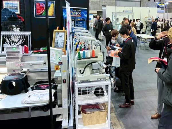 ASM OSAKAの展示ブースに並ぶ刺繍機と、機器を見学する来場者。